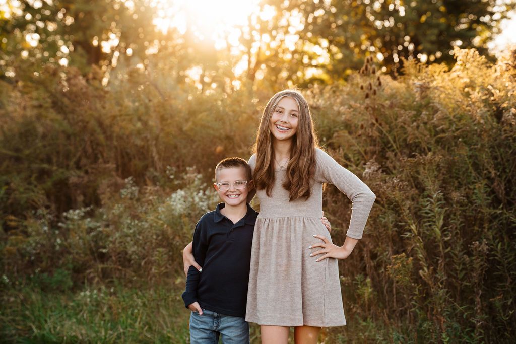 Two smiling children, a boy and girl, pose outdoors in golden sunset light surrounded by lush green foliage.