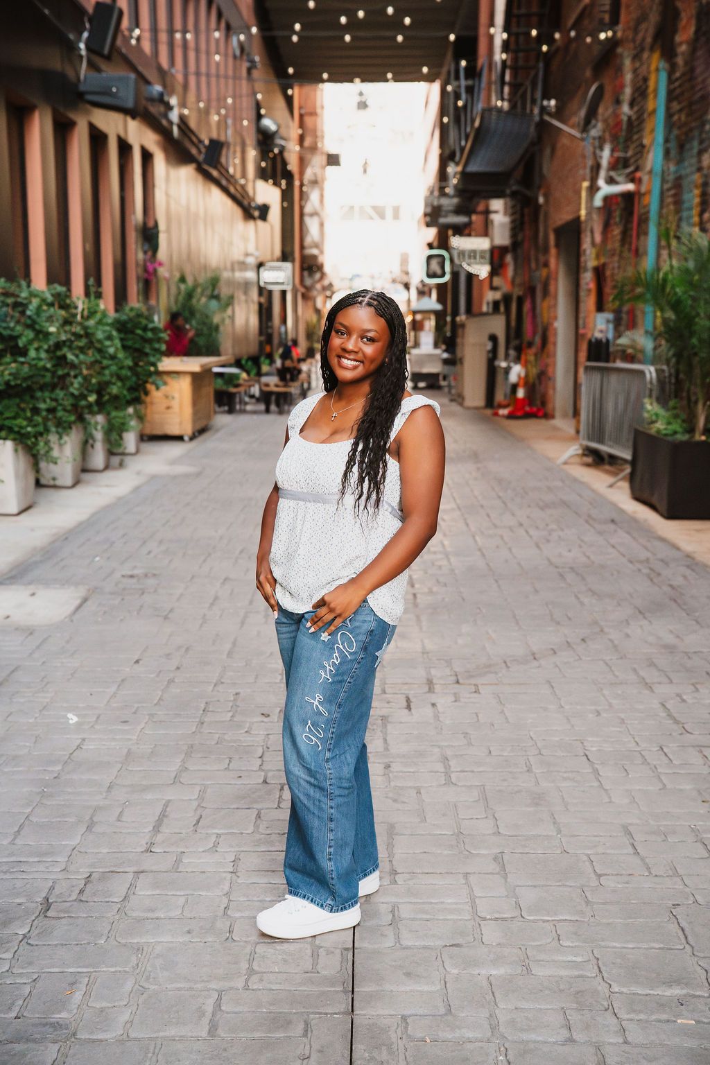 A person stands on a charming brick alley with string lights overhead and potted plants lining the walls.