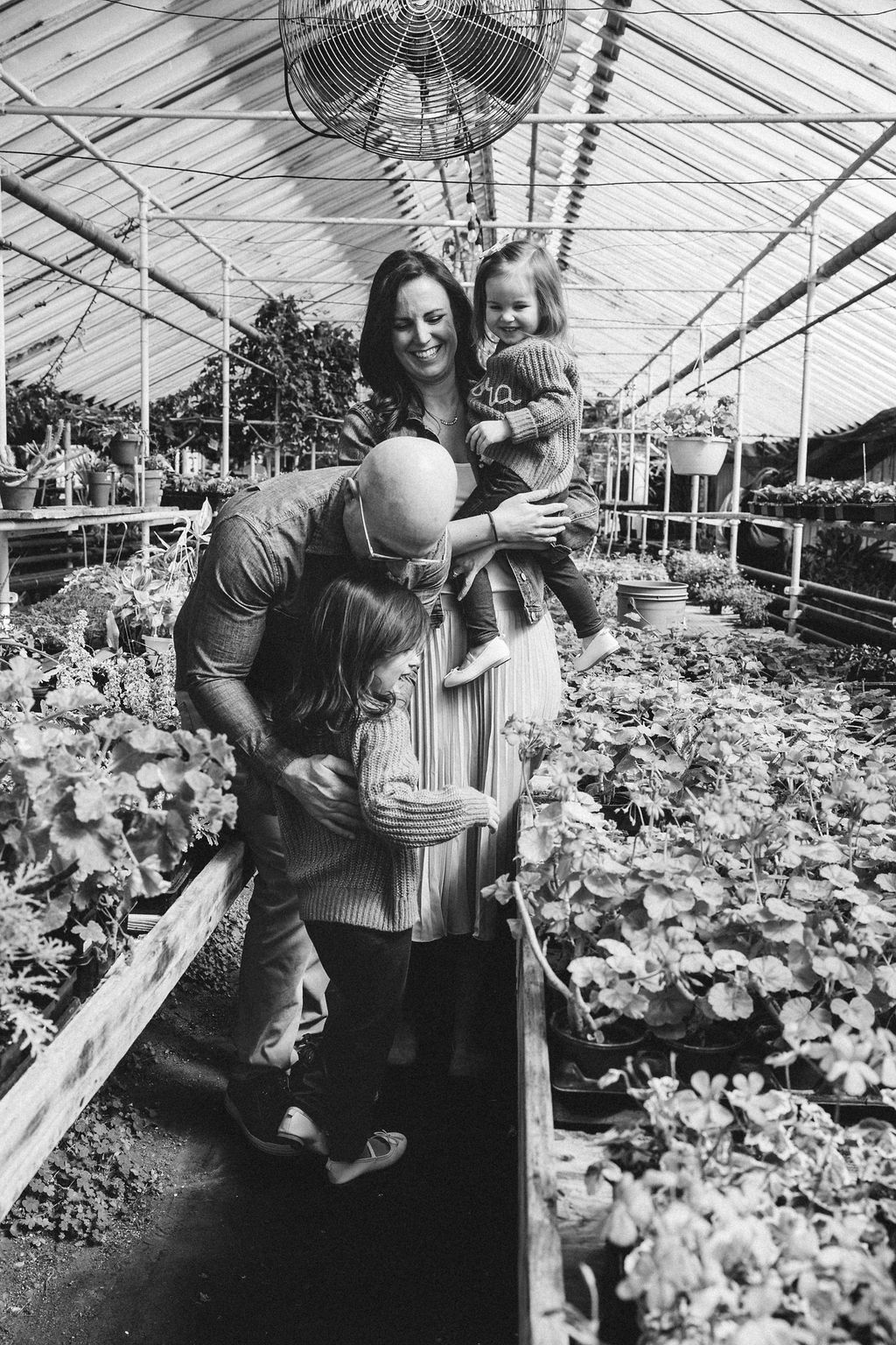 Black and white photo of a happy family with two children in a lush greenhouse filled with plants.