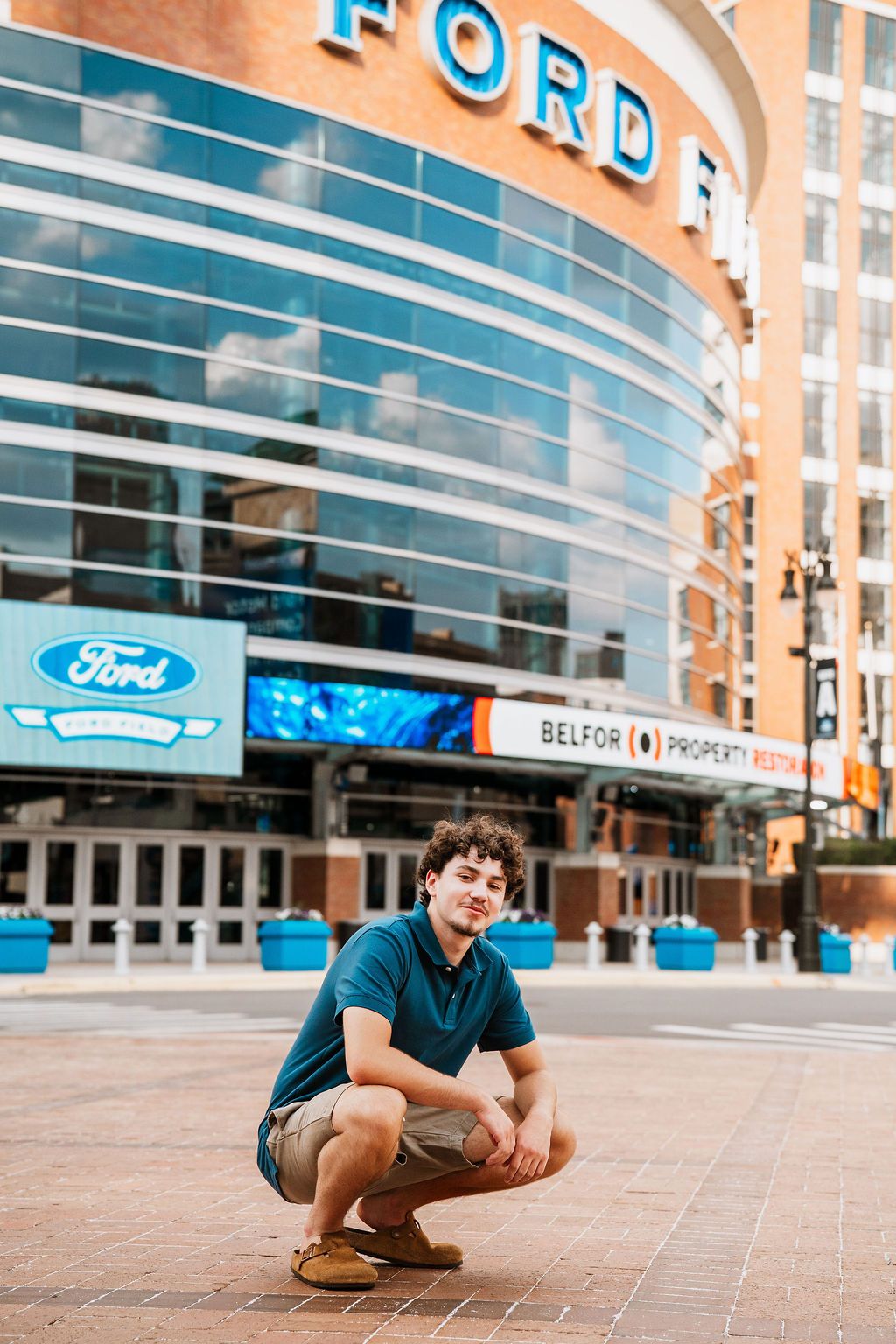 Young man crouching in front of Ford Field stadium in Detroit, wearing a teal polo shirt and khaki shorts.