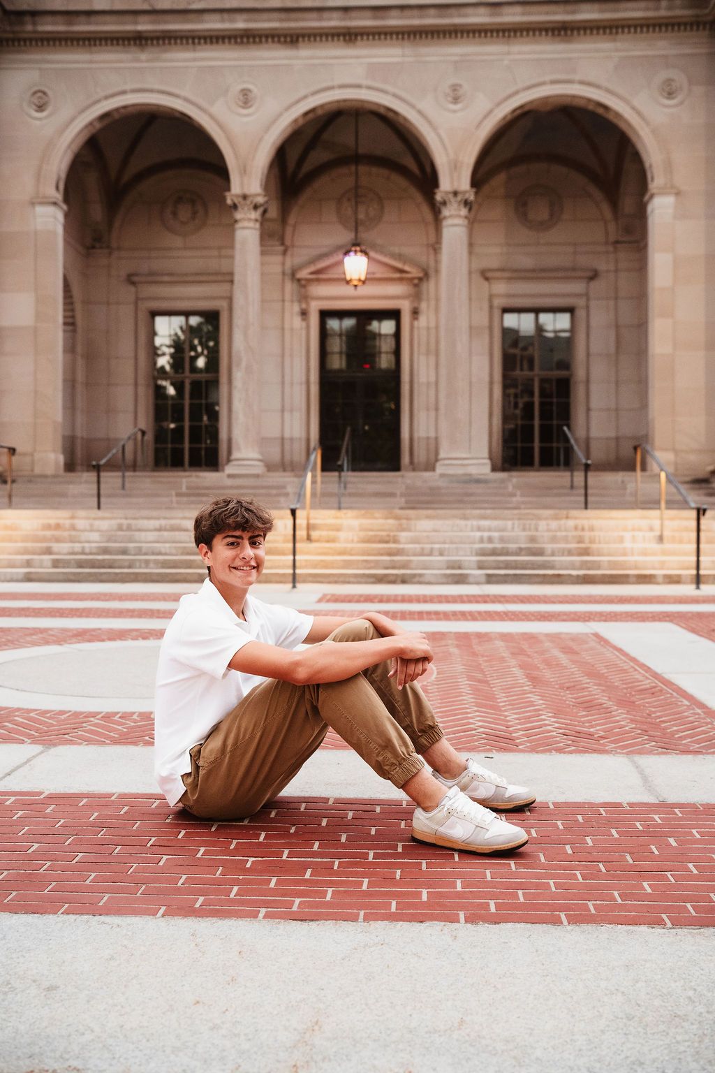 Young man sitting on brick plaza steps smiling in white tee and khakis before a grand neoclassical building.