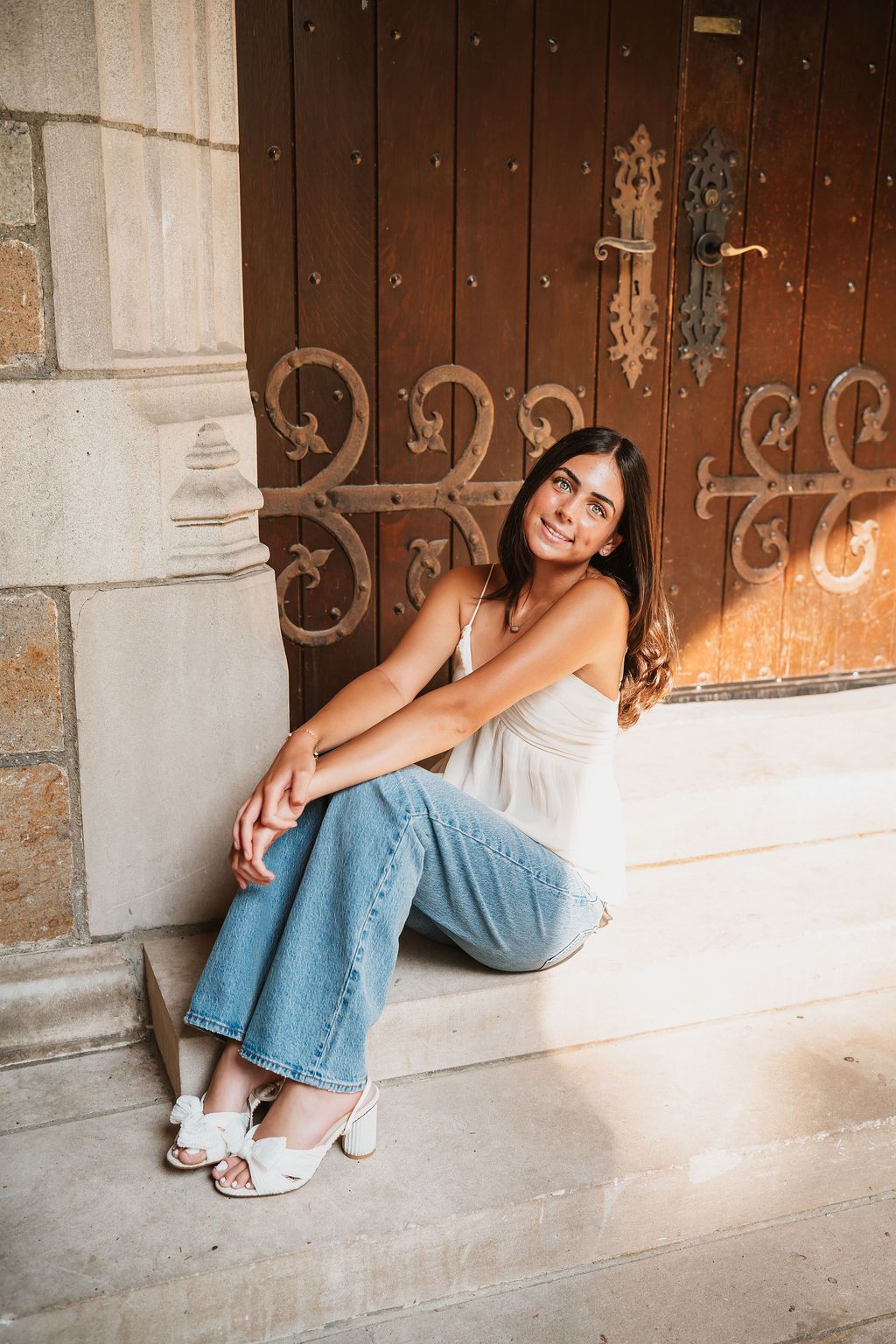 A person in jeans sits casually against a rustic wooden door with decorative metal scrollwork in an outdoor setting.