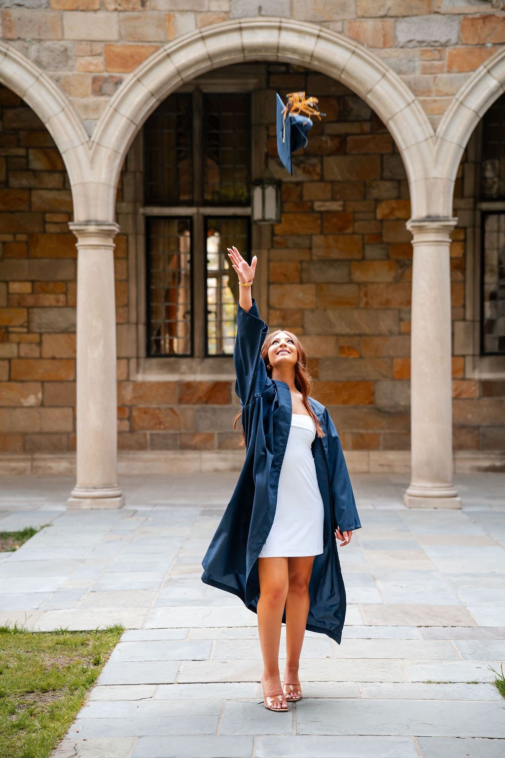 A person in a navy blazer and white dress celebrates joyfully under stone archways at a university campus.
