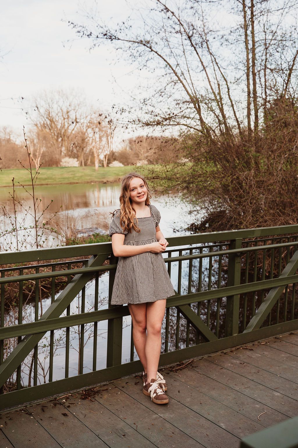 Young girl in striped dress smiling on a wooden bridge over a pond surrounded by trees.