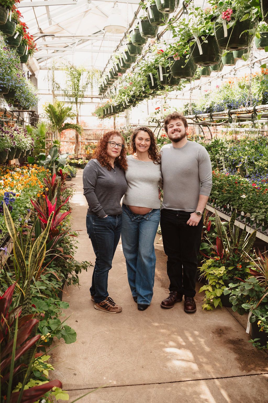 Three people smiling together in a lush greenhouse surrounded by colorful plants and flowers.