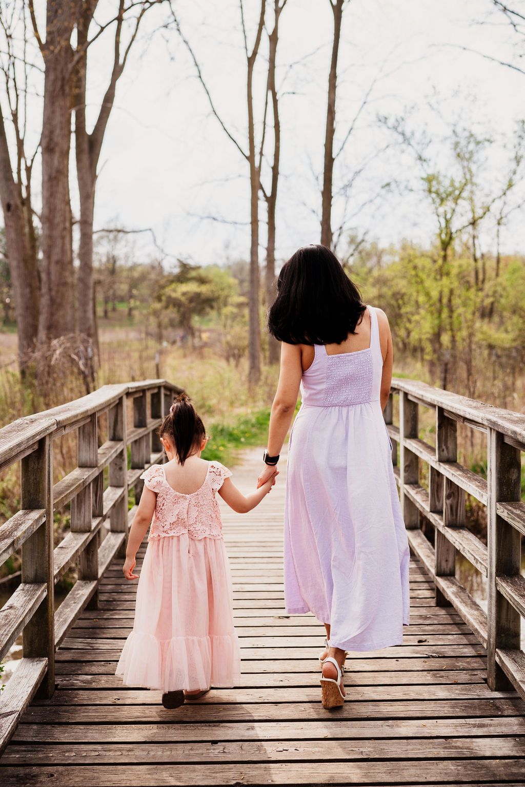 Two people in white and pink dresses walk hand in hand across a wooden boardwalk surrounded by bare trees.