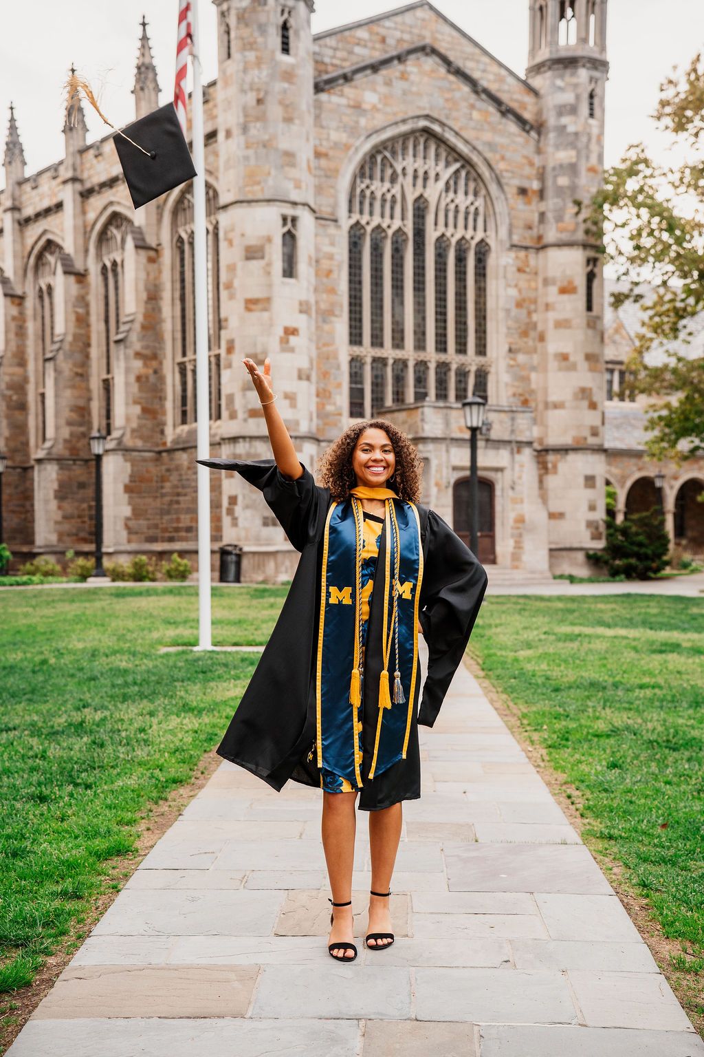 Graduate in blue and gold regalia celebrates joyfully in front of a Gothic cathedral-style university building.