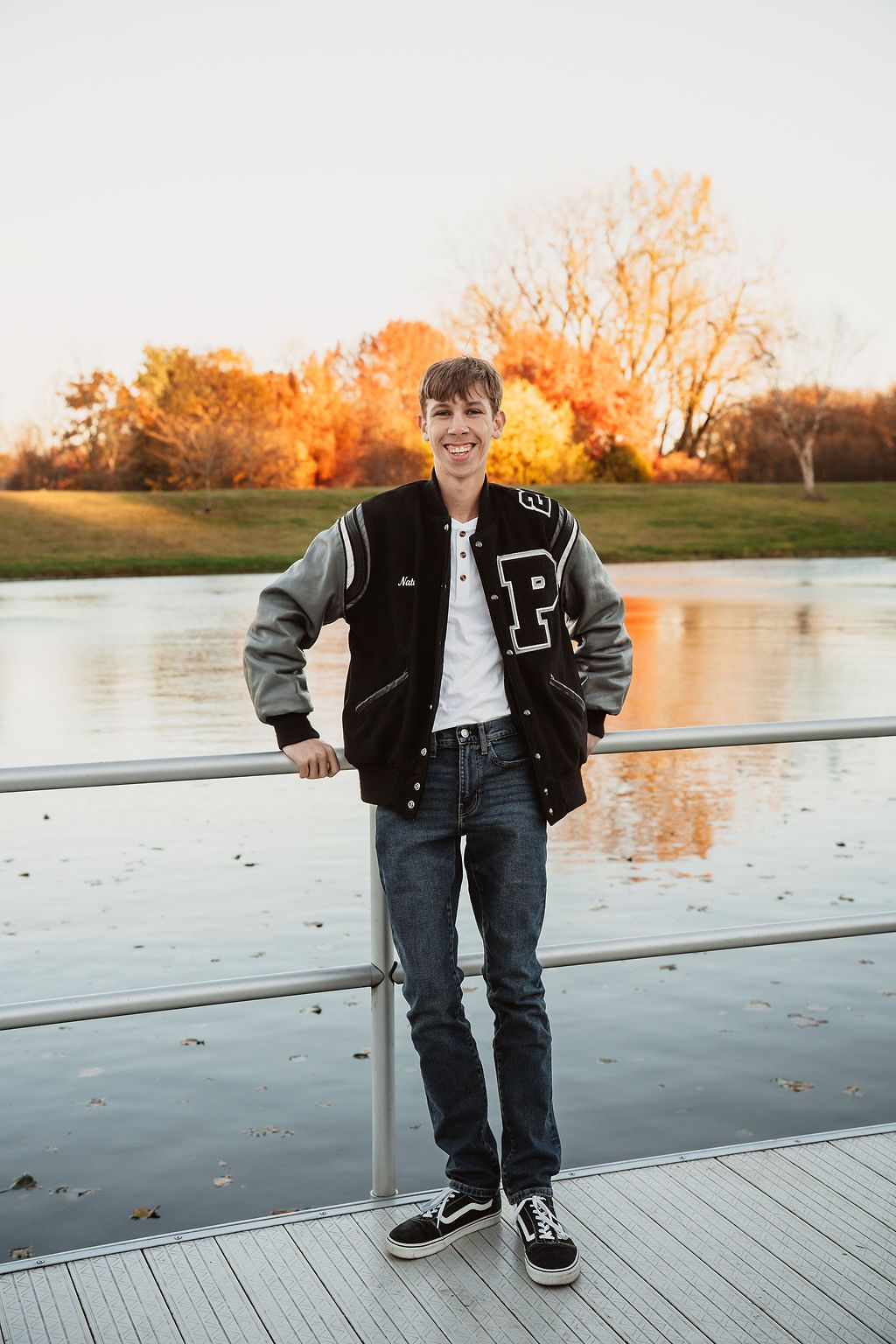 Smiling teen in varsity jacket standing on dock by lake with fall foliage senior photo.