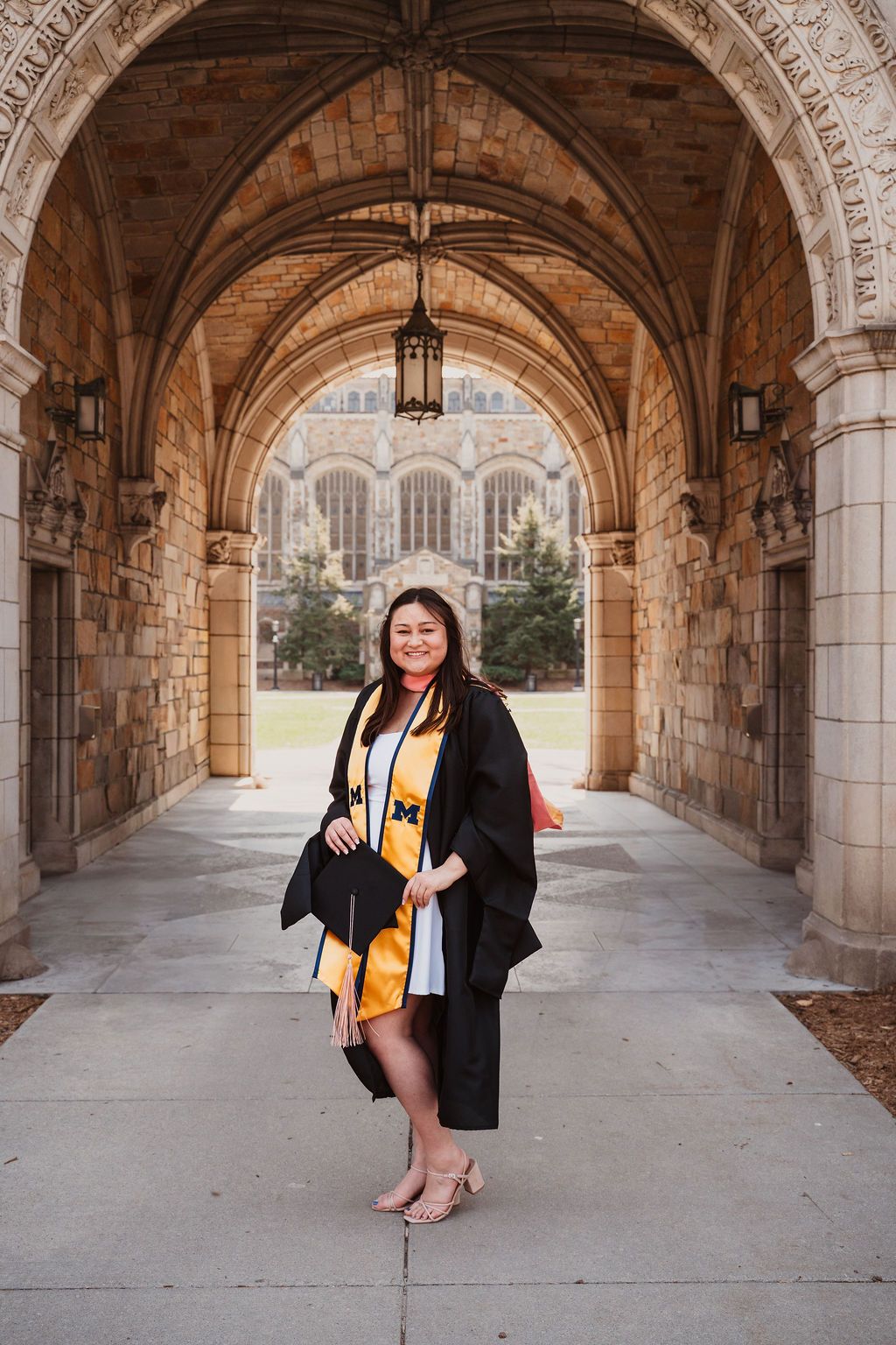 Graduate in black cap and gown with gold stole poses under stone archway at University of Michigan.