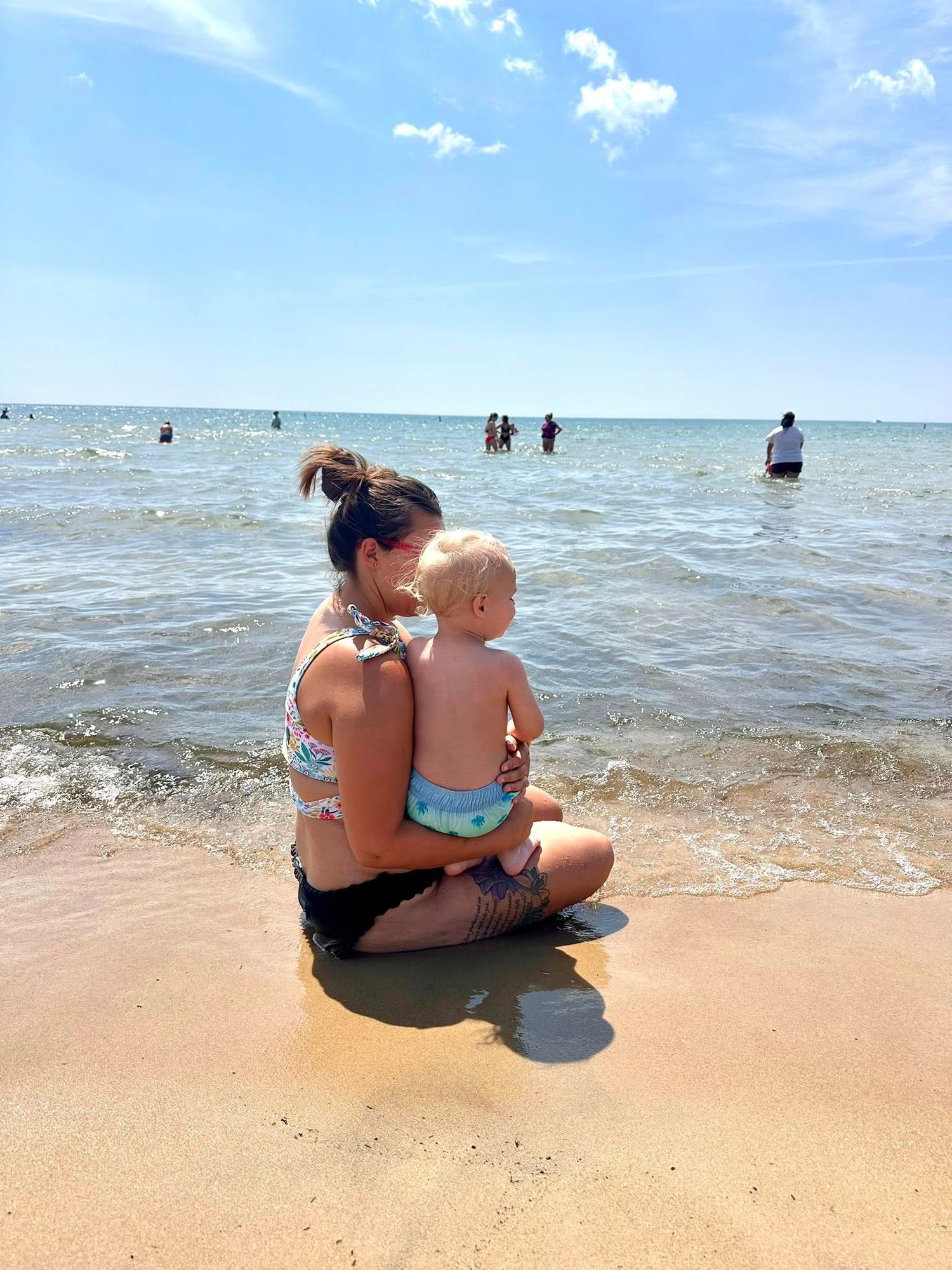 A mother sits on sandy beach shore holding her toddler while waves lap nearby and swimmers enjoy the ocean.