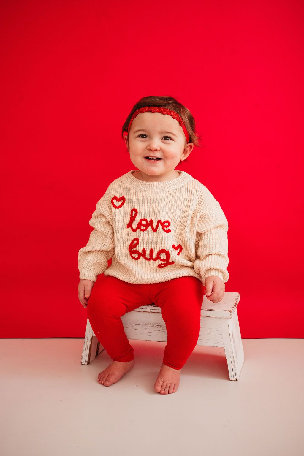 Smiling baby in cream love bug sweater and red pants sits on white stool against bright red background.