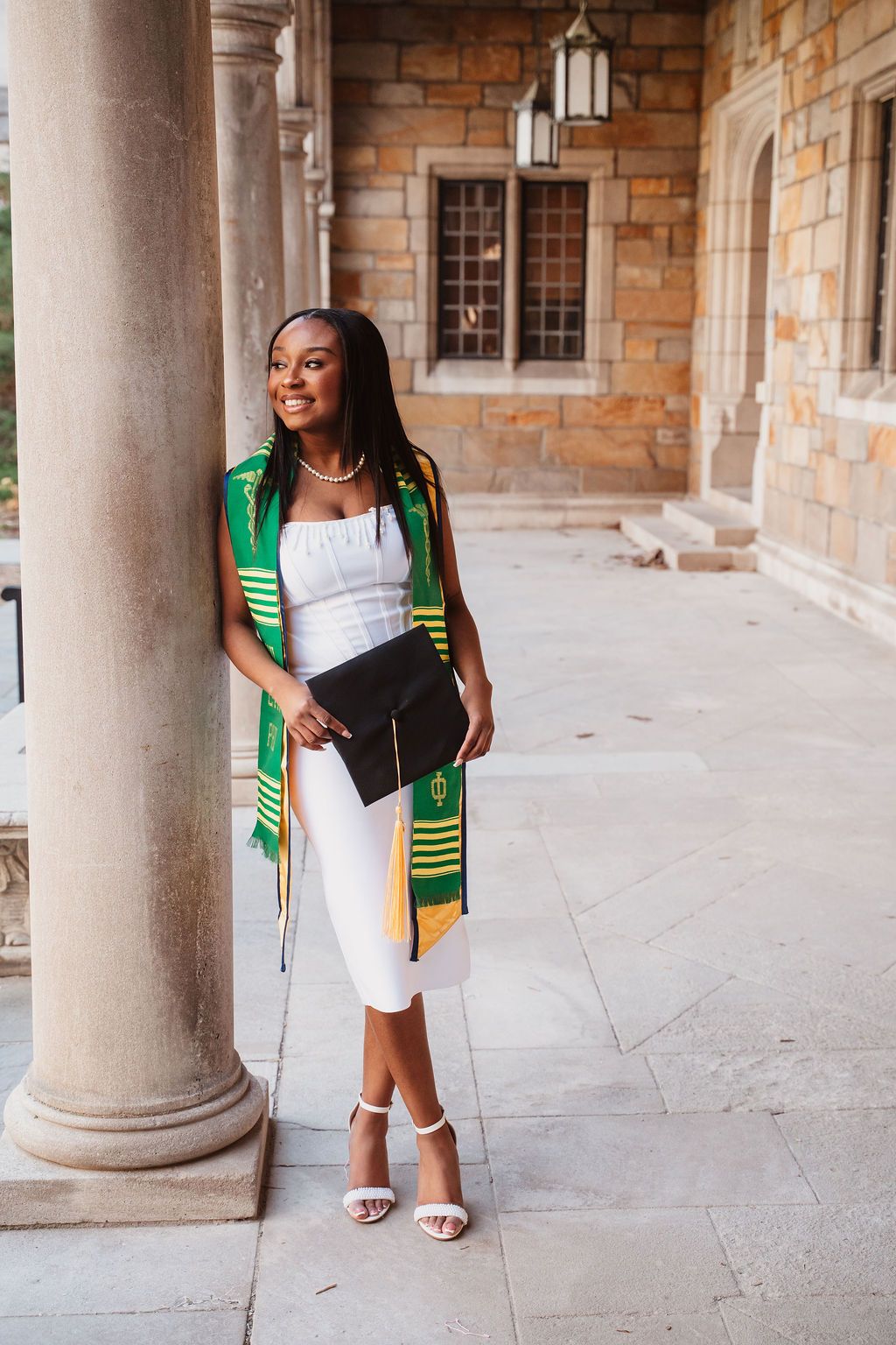 Young woman in white outfit wearing green and gold graduation stole, holding cap, posing by stone column.