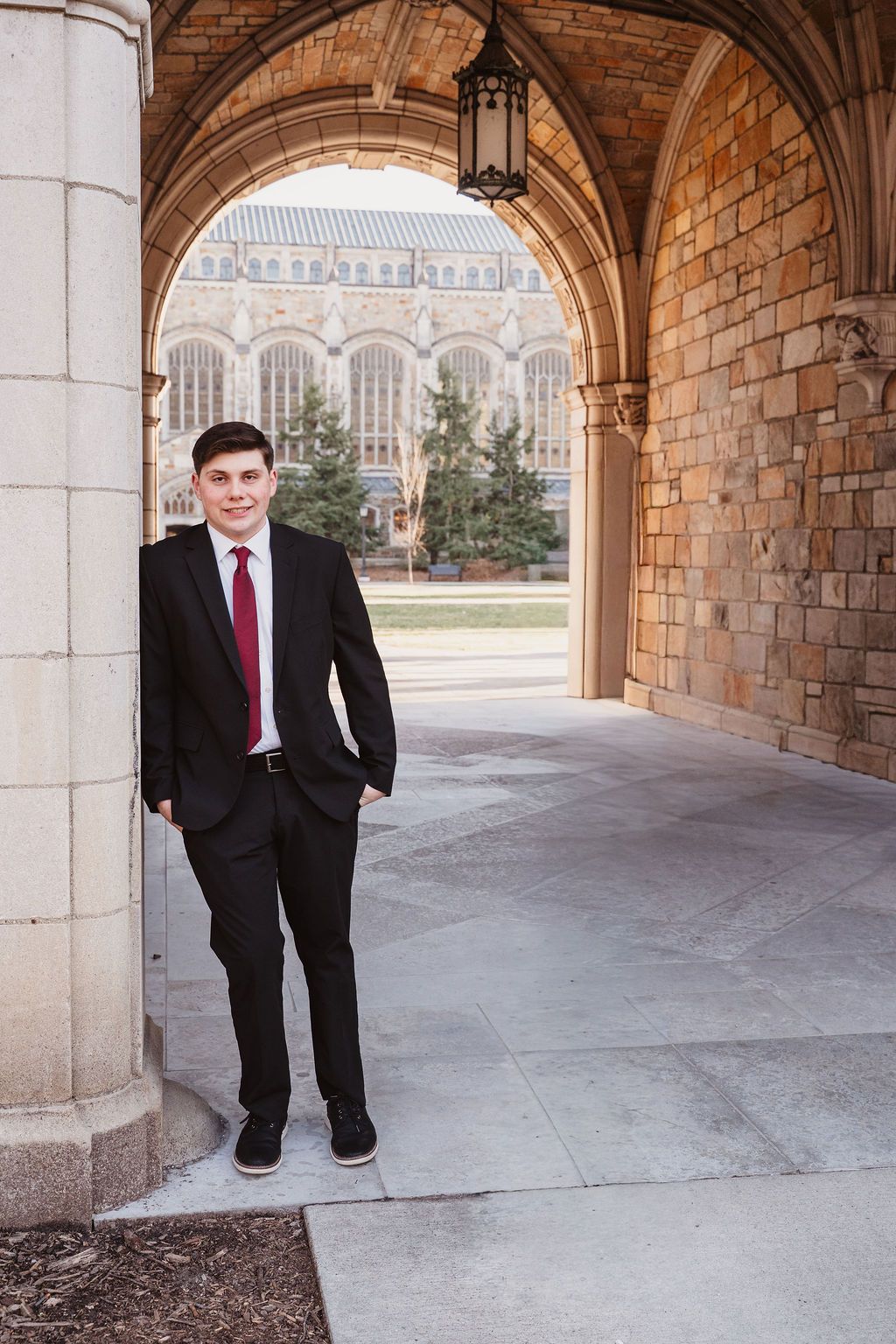 Young man in black suit and red tie posing under stone archway at university campus.