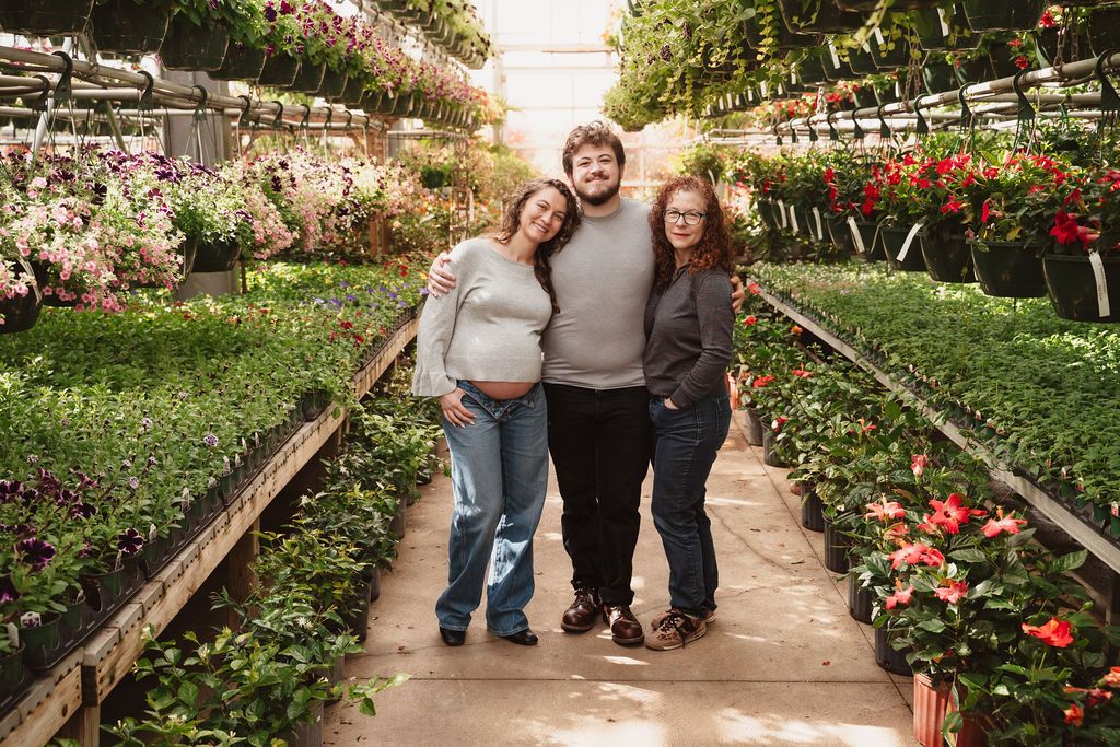 Three people smiling in a greenhouse nursery surrounded by colorful flowers and plants, one woman showing a baby bump.