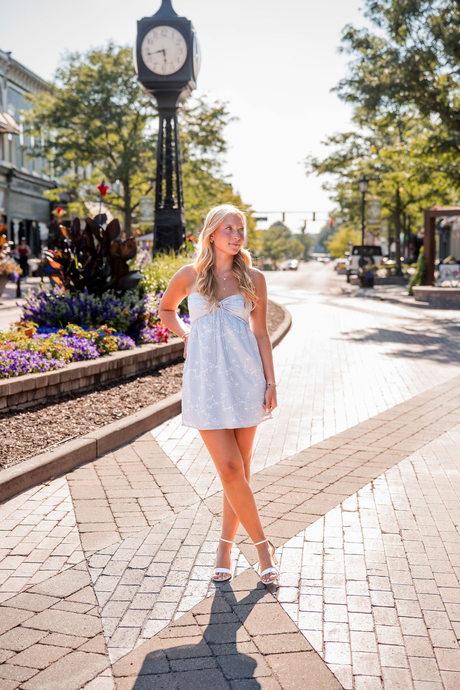 Blonde woman in white mini dress and heels poses on brick sidewalk near a street clock in a sunny downtown area.
