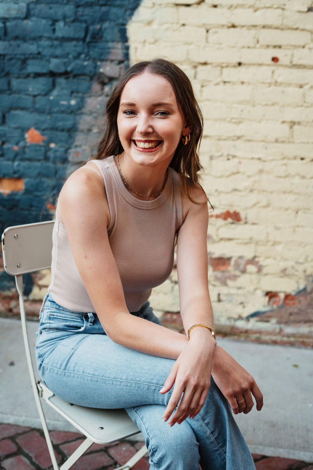 Smiling young woman sitting on a white folding chair outdoors, wearing a beige tank top and jeans against a brick wall.