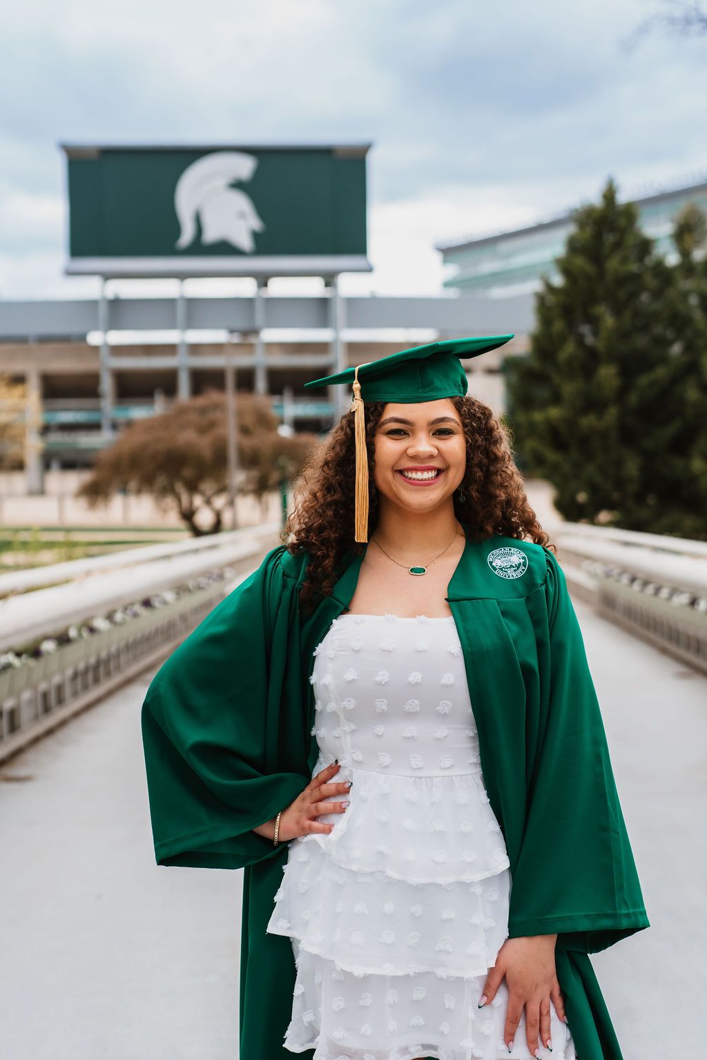 Graduate in green cap and gown poses for graduation photos on campus with Michigan State University signage.