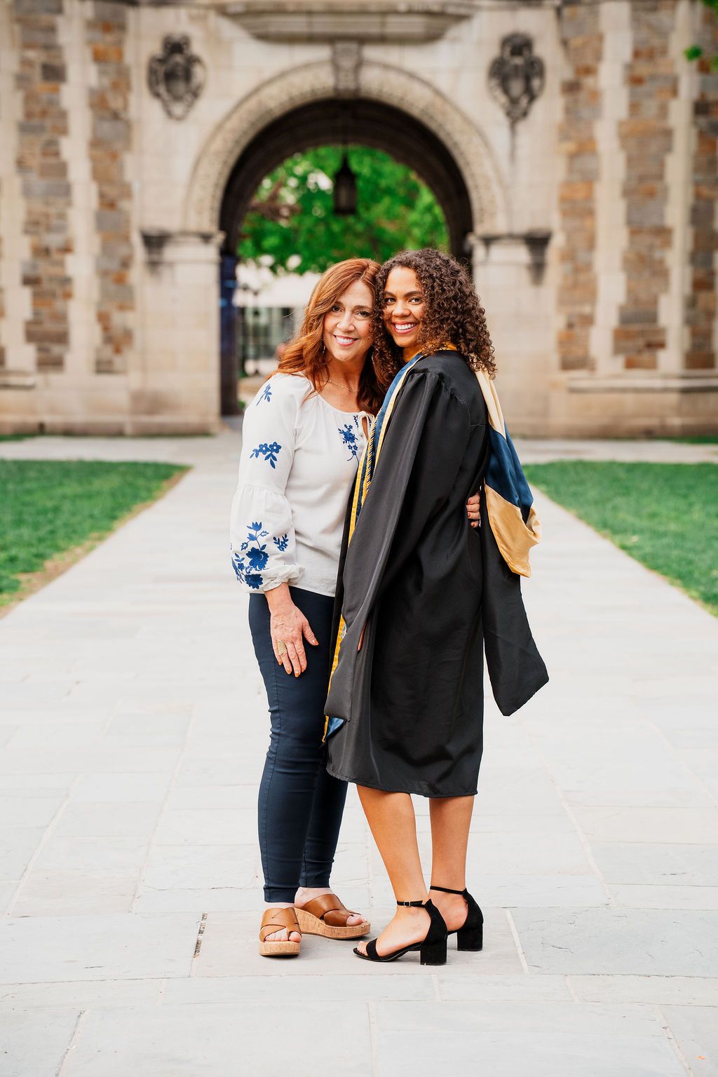 Two friends pose together on a college campus walkway with an ornate stone archway in the background.