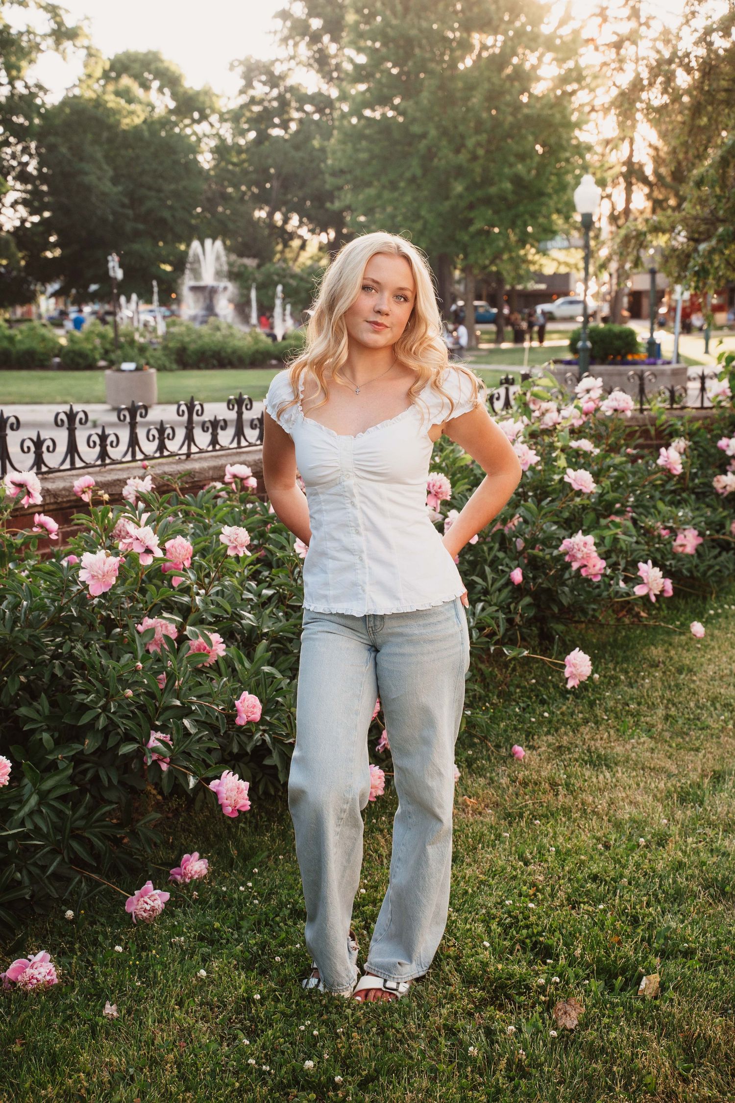 Blonde woman in white top and jeans posing by pink peonies in a park with a fountain backdrop.