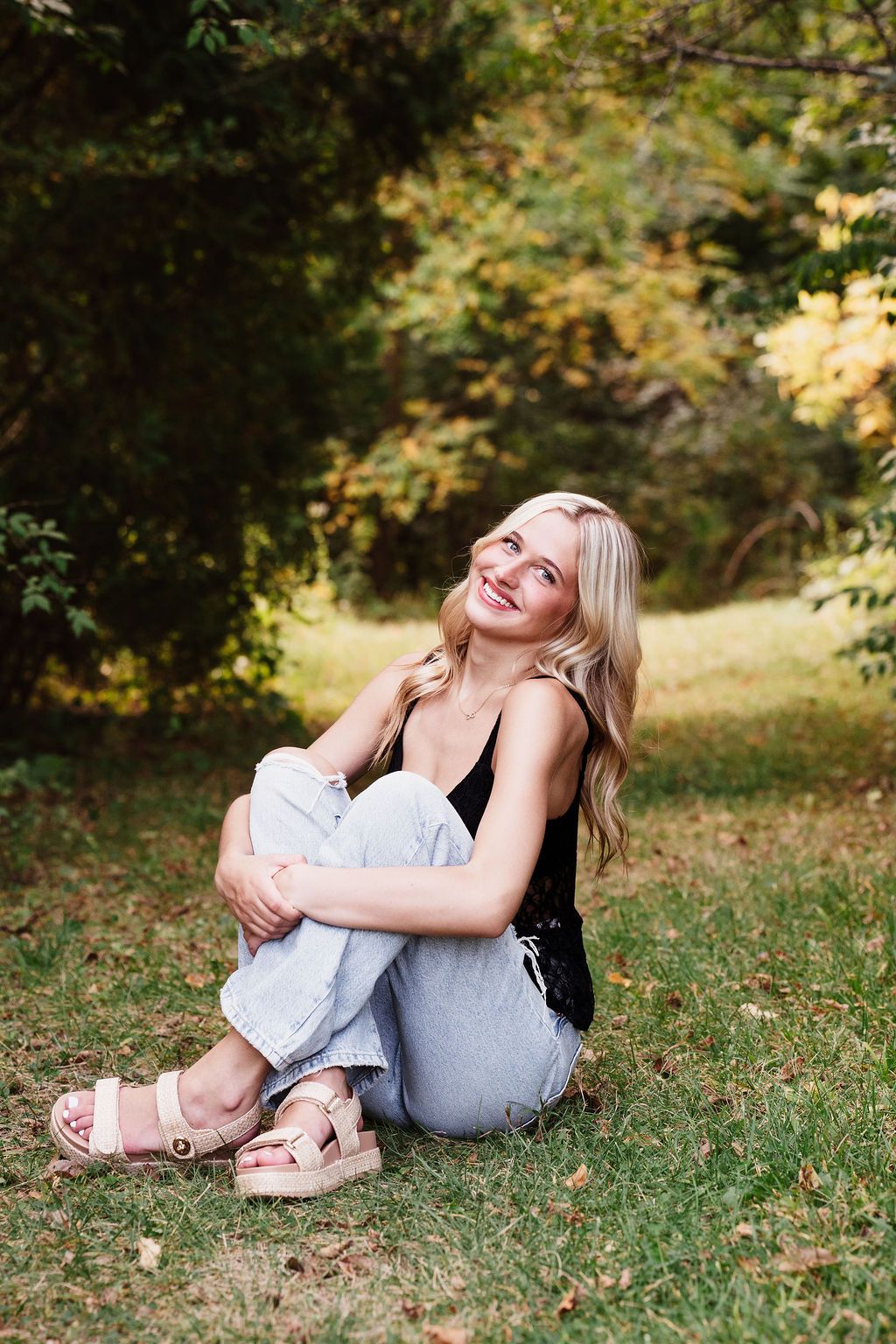 Smiling blonde woman in black top and jeans sitting on grass surrounded by lush green trees.