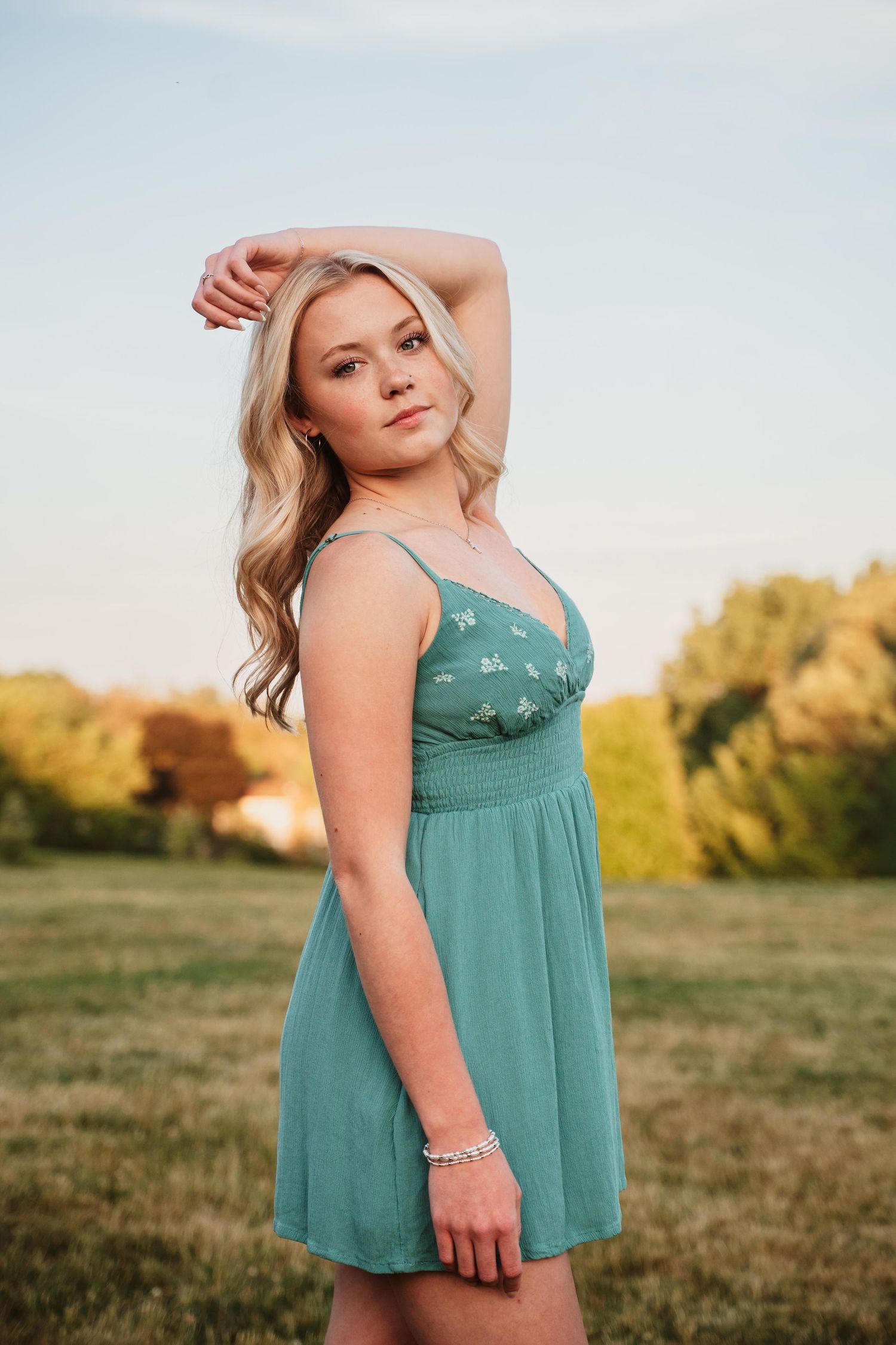 Young blonde woman in teal floral sundress posing outdoors in a green field during golden hour.