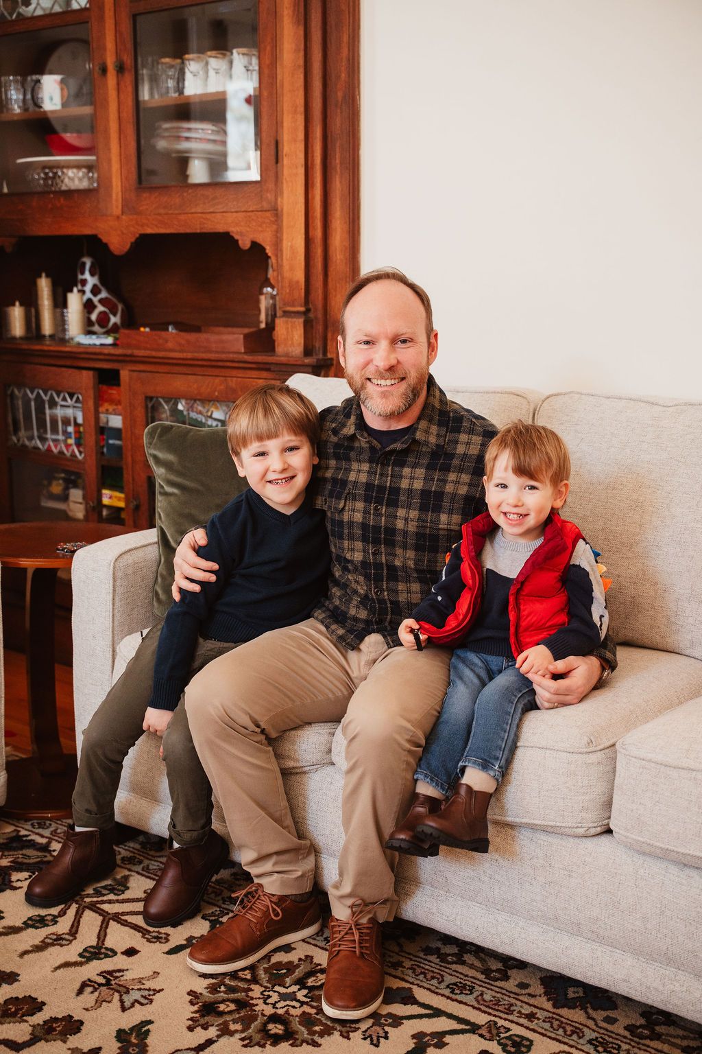 Father sitting on a couch smiling with two young boys in a cozy living room with wooden cabinet.