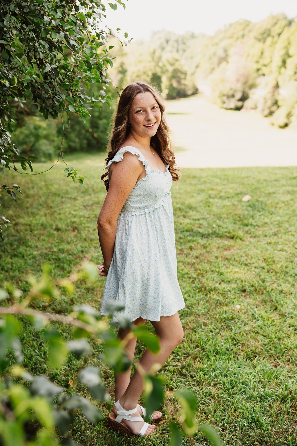 Smiling teenage girl in a light blue sundress standing on green grass near leafy tree branches outdoors.