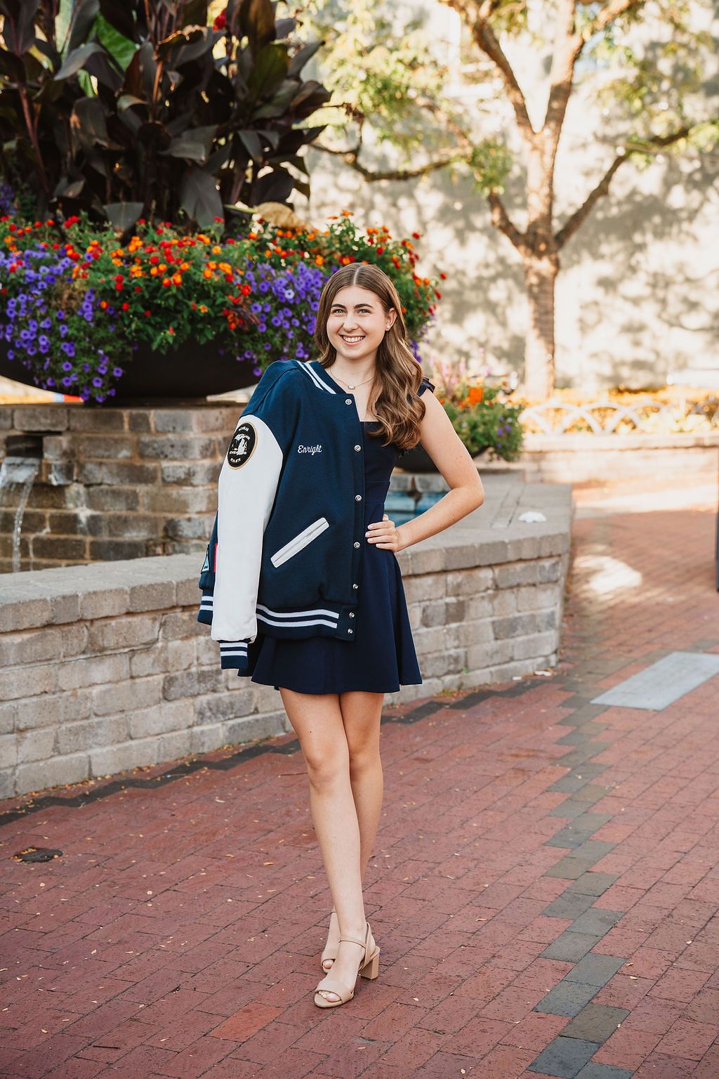 Young woman in navy dress holding varsity jacket poses on brick walkway with colorful flowers.