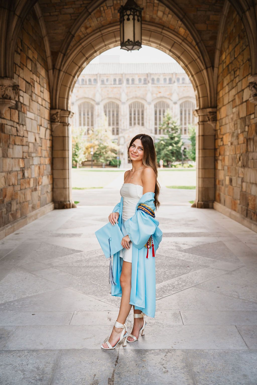 Woman in light blue dress poses gracefully under historic stone archways on university campus.