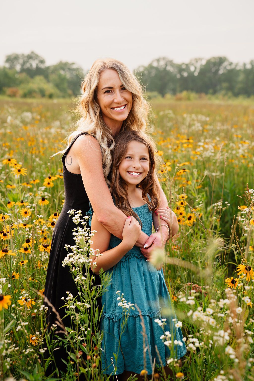 A smiling woman in black dress hugs a young girl in teal dress in a vibrant wildflower field.