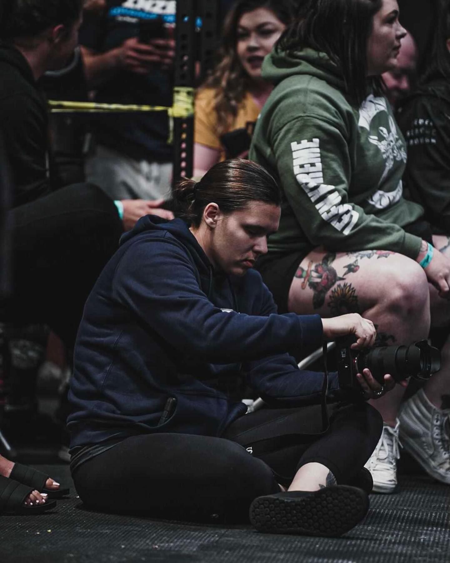 Woman in navy hoodie tattooing a person seated on the floor at an indoor event.