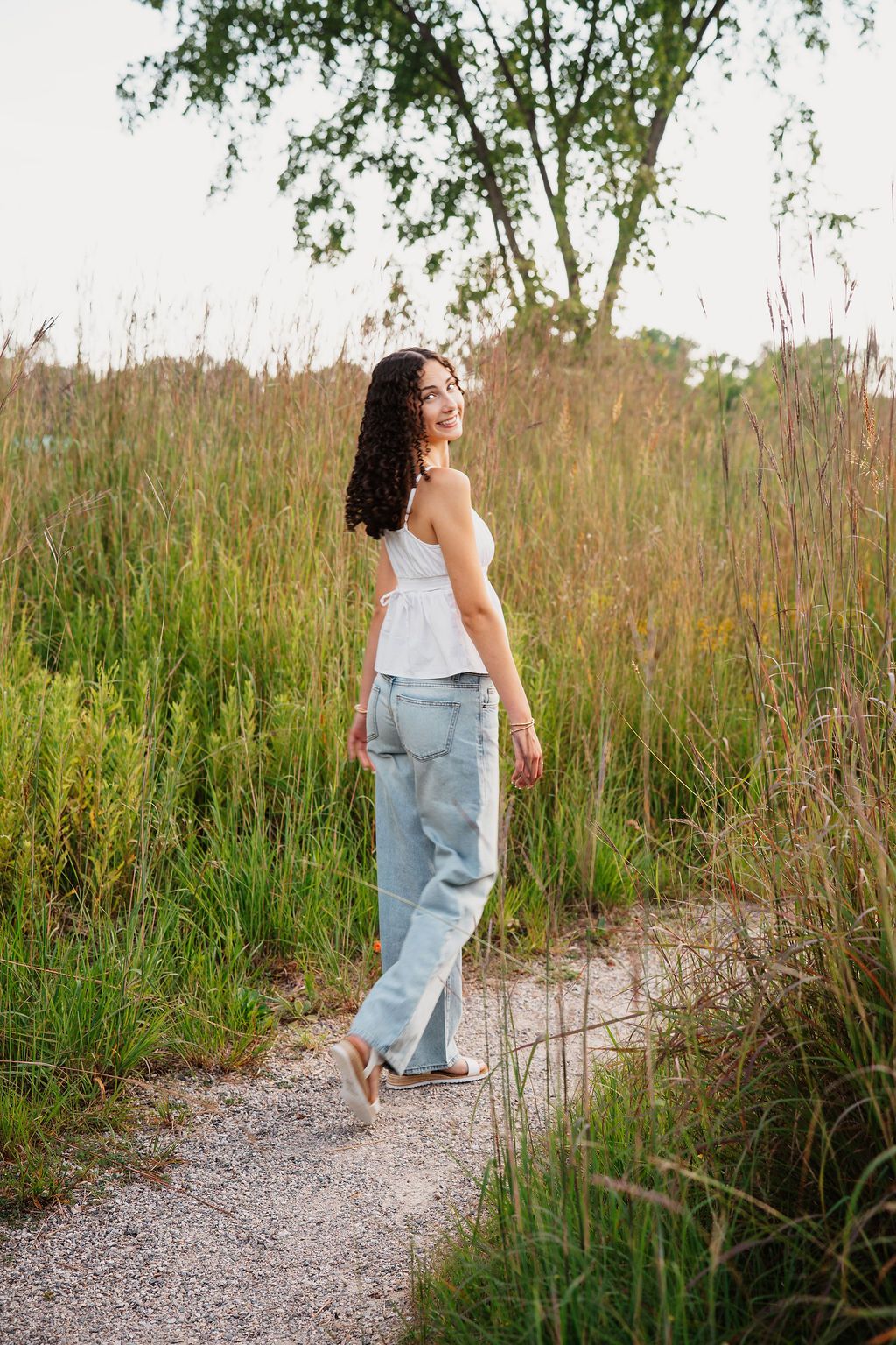 A person in a light blue dress walks along a gravel path surrounded by tall coastal grass and natural vegetation.