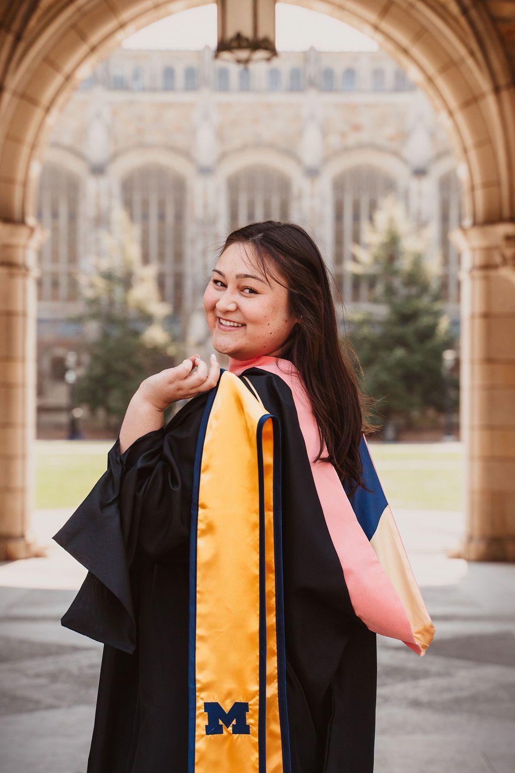 Graduate in black gown with maize Michigan stole poses under Gothic arch on U of M campus.