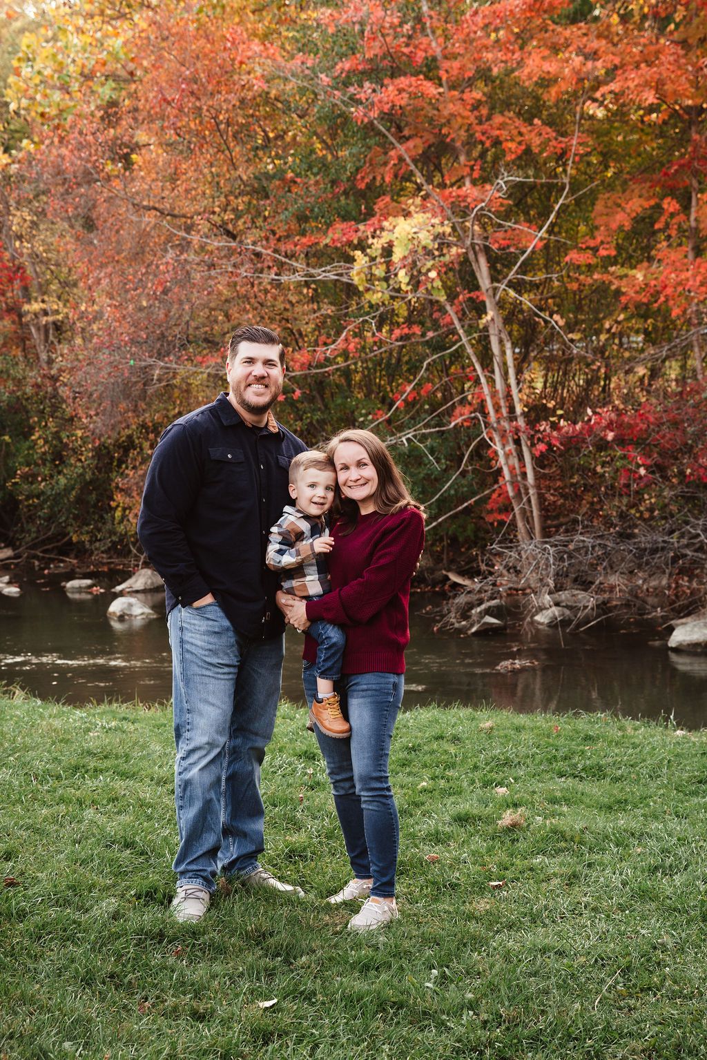 Family of three posing outdoors with vibrant red and orange fall foliage and creek in background.