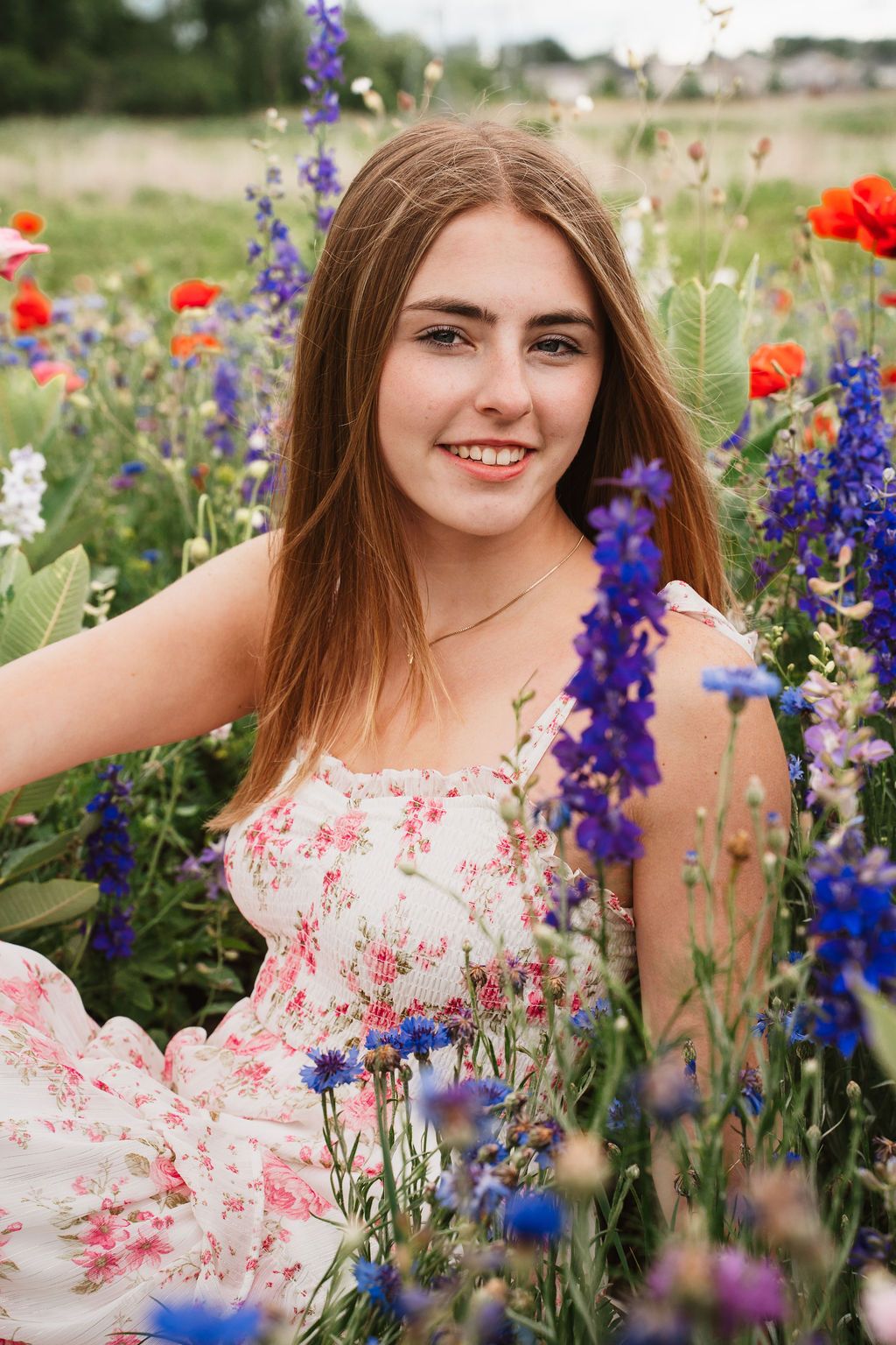 Young woman in floral dress smiling among colorful wildflowers including poppies and cornflowers in a lush field.