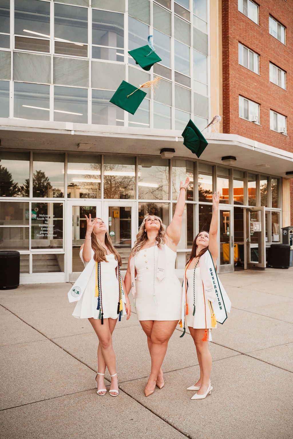 Three graduates in white dresses toss green caps in the air outside a brick university building.