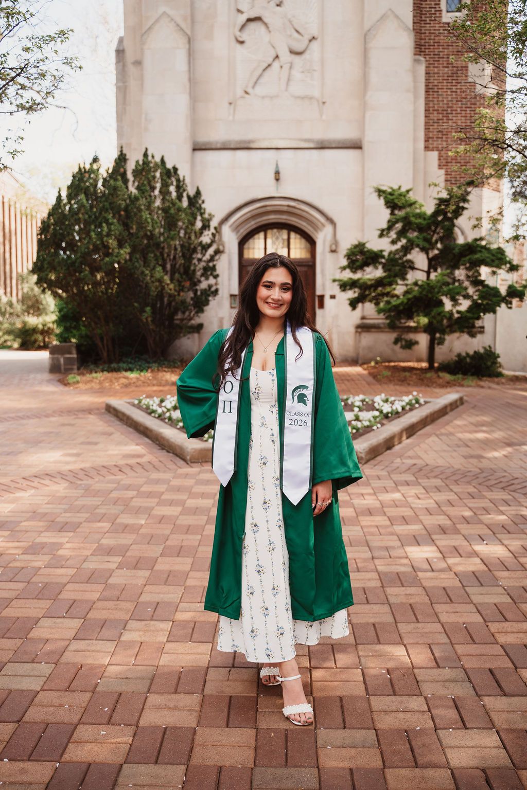 Young woman in green Michigan State University graduation cap and gown posing on brick plaza outside campus building.