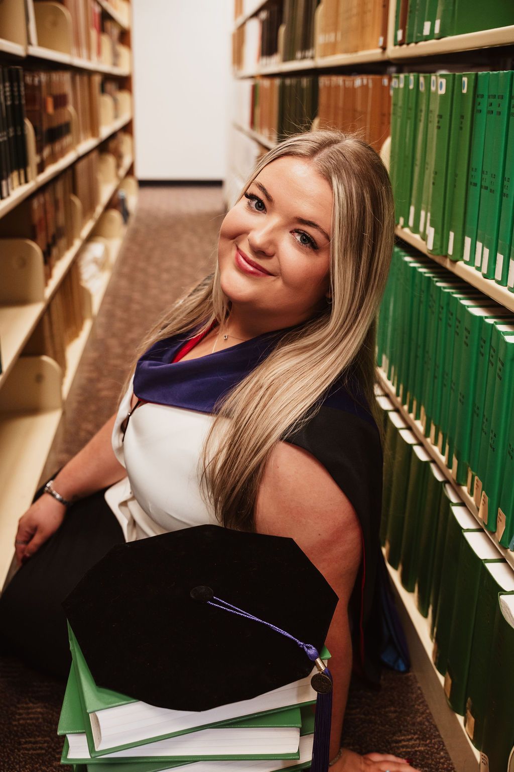 Smiling blonde woman in graduation gown sitting between library shelves holding mortarboard cap and green books.