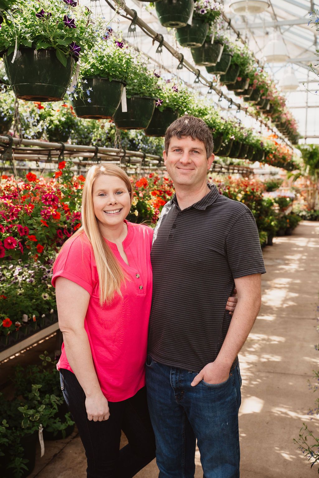 Smiling couple posing together in a lush greenhouse surrounded by colorful hanging flower baskets and blooming plants.