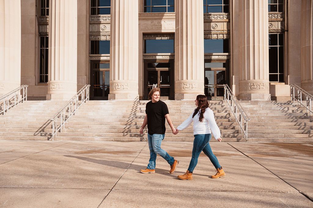 Couple holding hands walking in front of a grand building with tall columns on a sunny day.