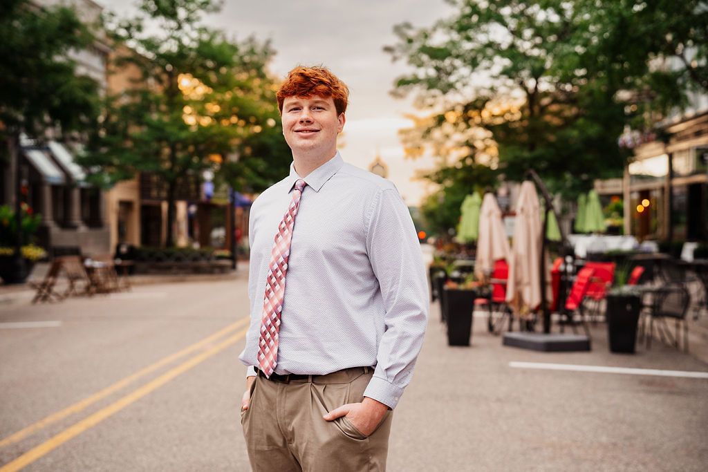 Young man with red hair wearing a dress shirt and tie stands confidently on a street in an outdoor dining district.