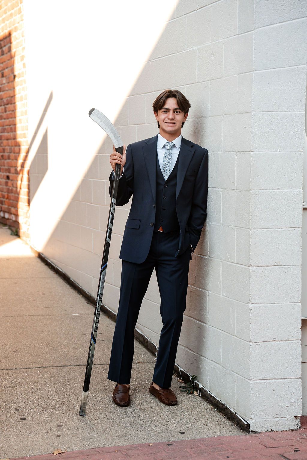 Young man in navy suit holding hockey stick leaning against brick wall outdoors.