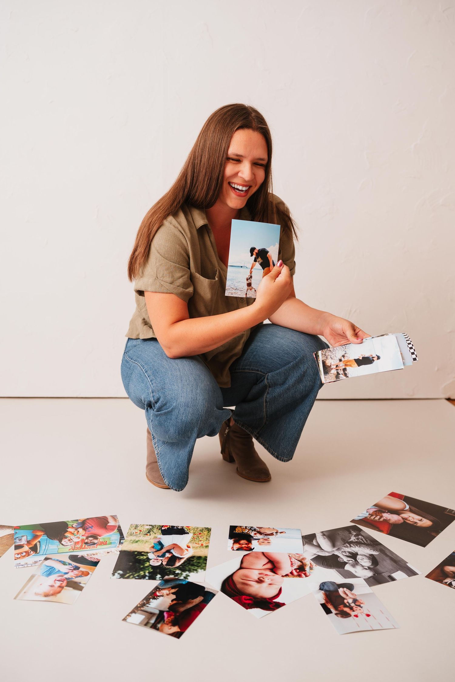 Smiling woman in an olive shirt and jeans crouches on a white floor holding printed photos, with more prints spread out around her.