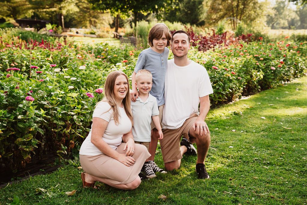 Family of four posing in a lush garden with colorful flowers, smiling outdoors on a sunny day.