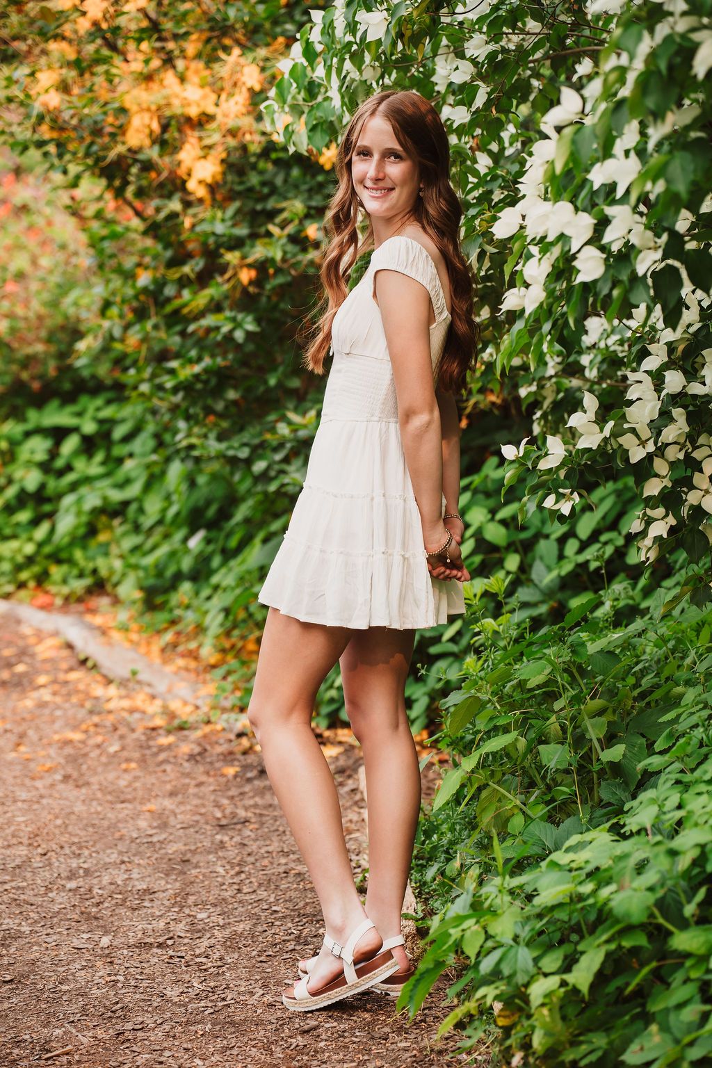 Young woman in white mini dress and sandals smiling on garden path surrounded by lush green foliage and white flowers.