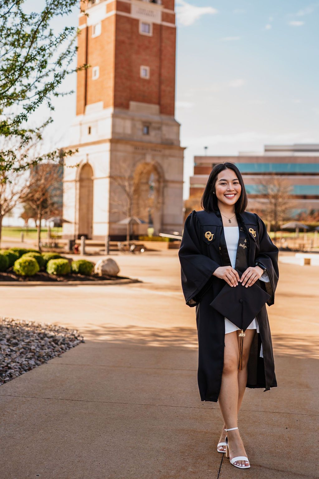 A graduate in a black gown poses near a brick building on a sunny day for a series of celebratory photos.