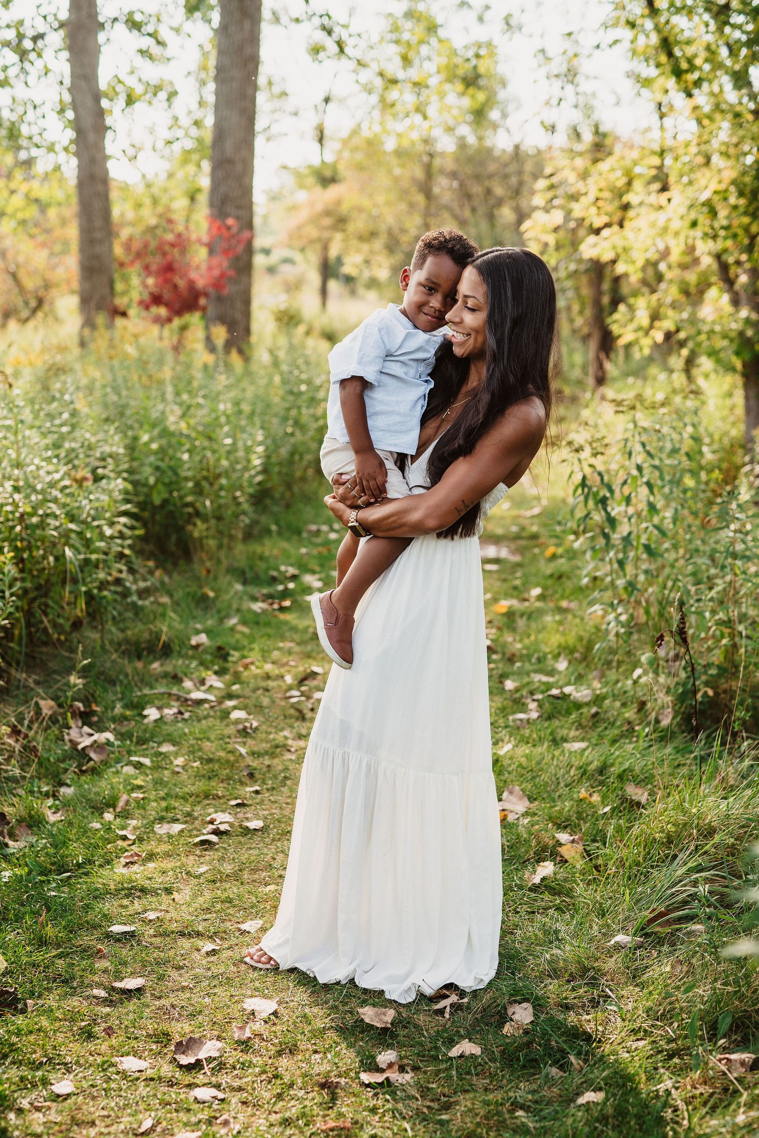 A tender outdoor family photoshoot in a sunlit wooded area with white dress against natural greenery.