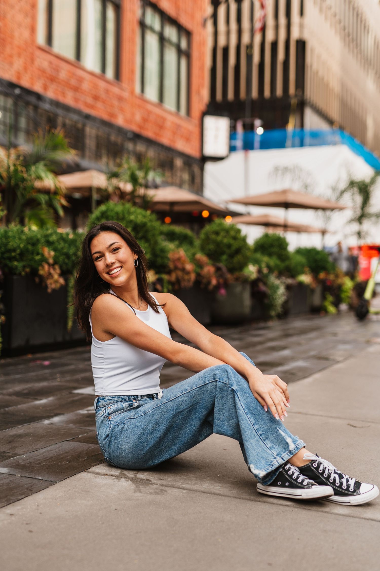 Smiling young woman sitting on urban sidewalk wearing white tank top, blue jeans, and black Converse sneakers.