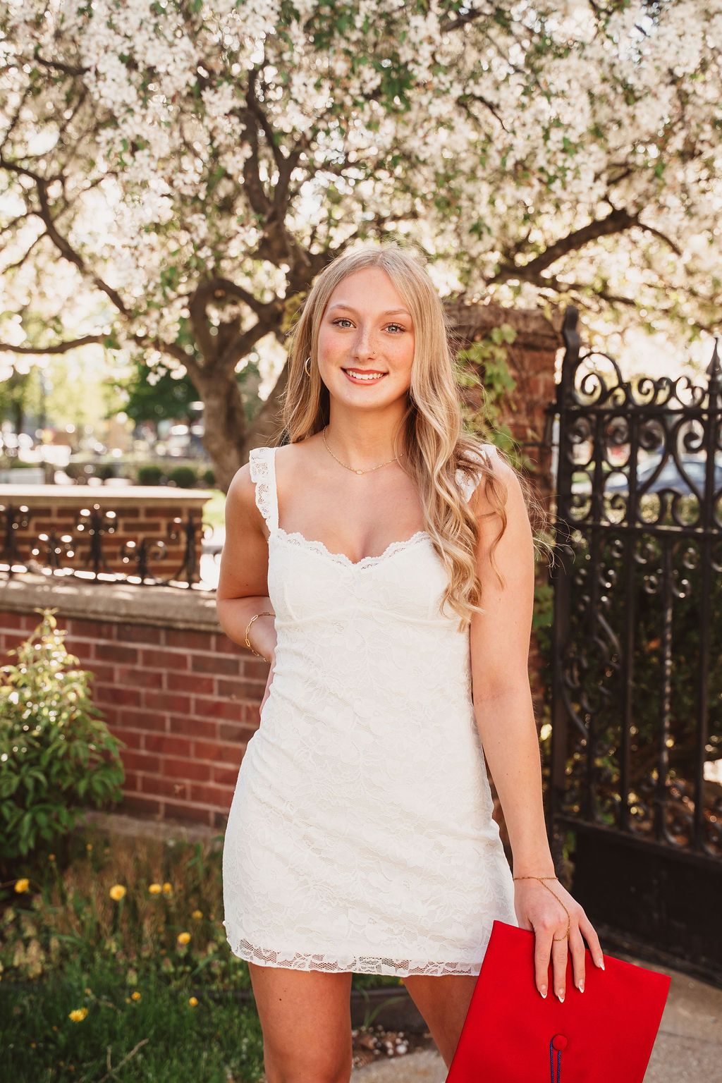 Young woman in white lace mini dress holding red folder, posing by flowering tree and brick wall.
