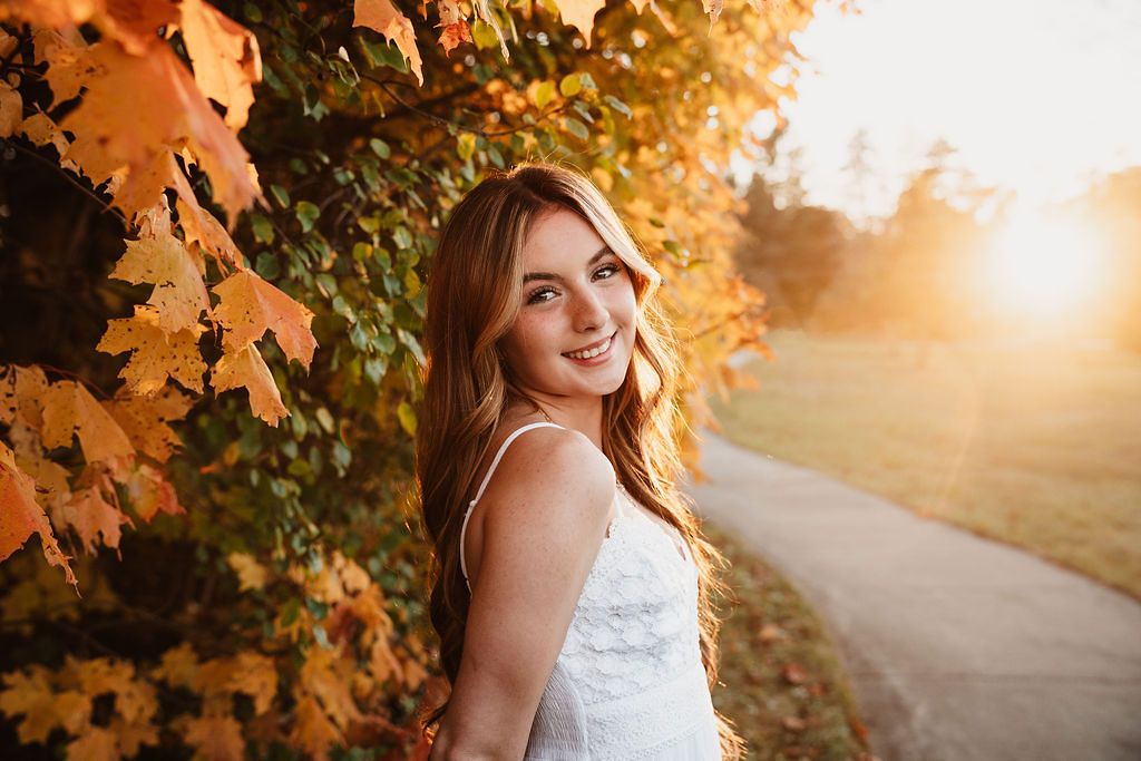 Young woman in white dress smiling outdoors among autumn foliage with golden sunset light in background.